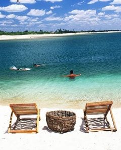 imagem de uma das praias em Fortaleza, com duas cadeiras de madeira, um cesto trançado e três pessoas mergulhando em um mar ciano