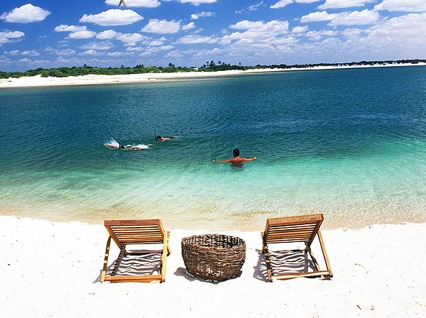 imagem de uma das praias em Fortaleza, com duas cadeiras de madeira, um cesto trançado e três pessoas mergulhando em um mar ciano
