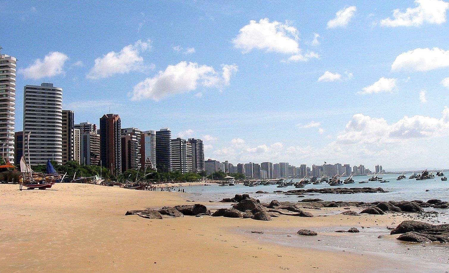 imagem de outra das praias em Fortaleza, a praia do Mucuripe. A imagem mostra a areia, as pedras, a rala vegetação e os prédios na orla da praia.