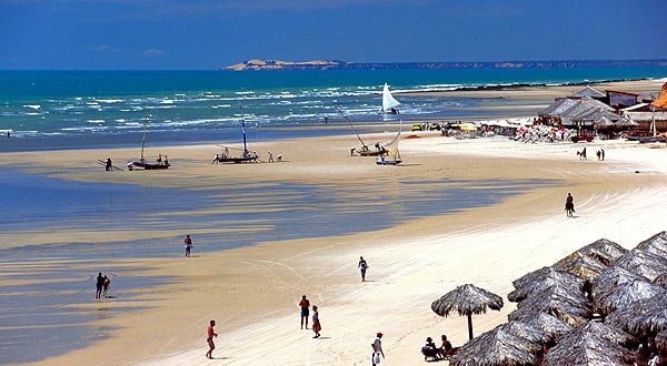 imagem de Canoa Quebrada, praia em Fortaleza. a imagem mostra o céu azulado e o mar, a areia e algumas pessoas e barcos