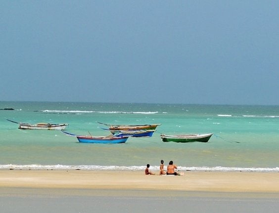 Uma das praias em Fortaleza, de maré baixa, é a praia do Mudaú, presente na imagem de céu azul, cinco barcos e três pessoas na beira do mar