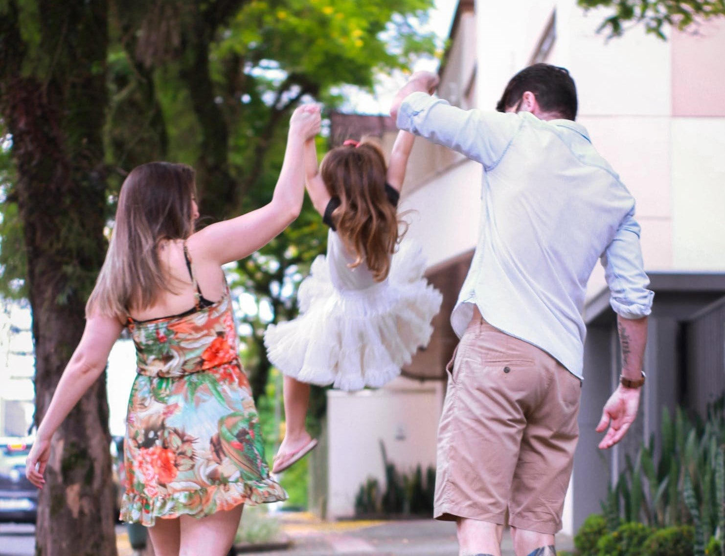 Mulher e homem segurando e levantando pelas mãos uma criança de vestido que está indo passear.