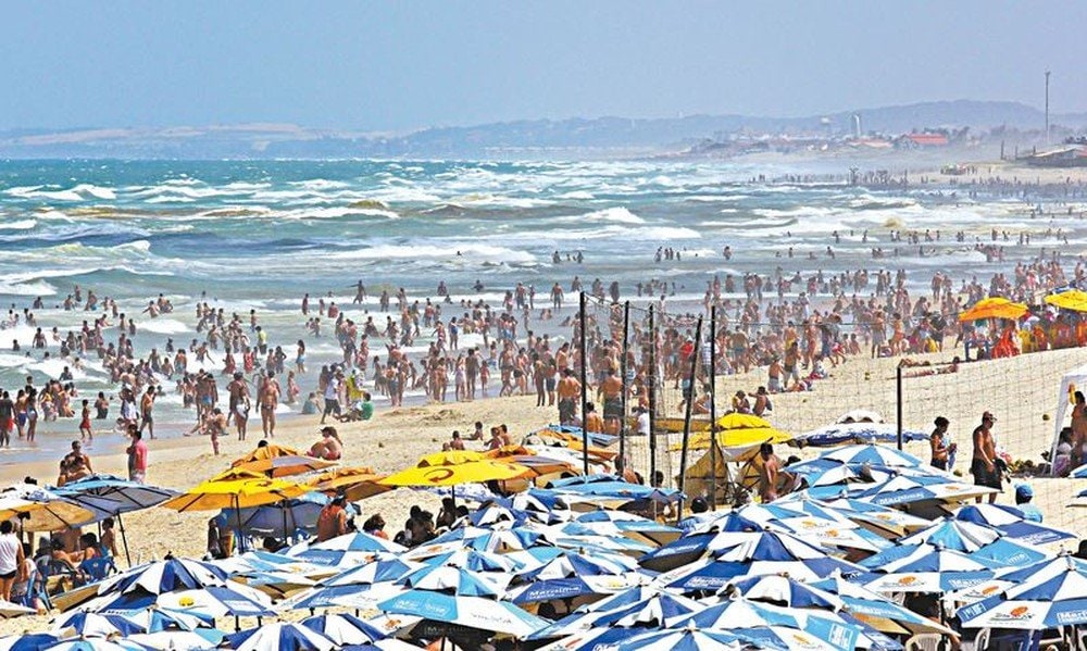 imagem de uma das melhores praias de Fortaleza, a Praia do Futuro. Na imagem, há milhares de pessoas e centenas de guarda sois, com areia, mar e montanhas.